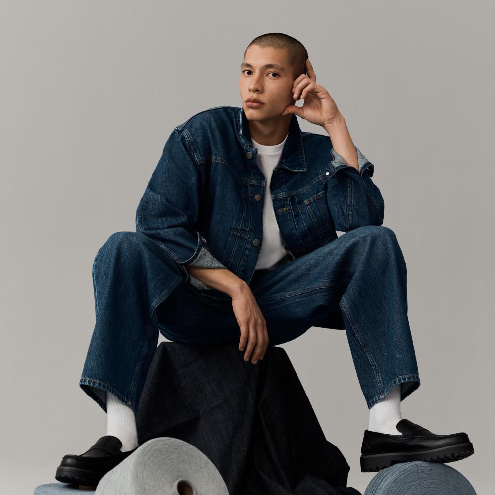 Person wearing denim jacket and jeans with white T-shirt, sitting on a stool in a thoughtful pose.