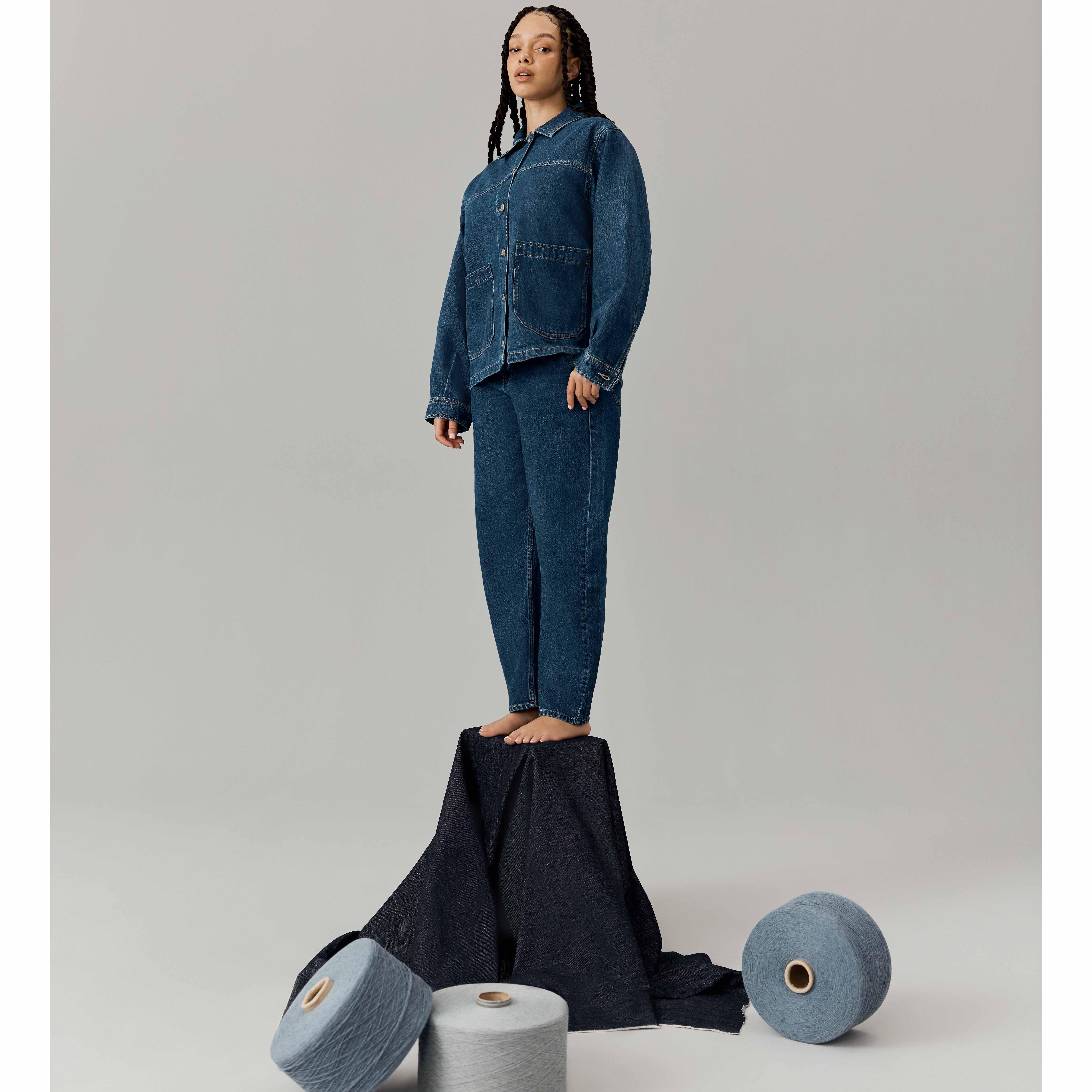 A person in blue denim clothes stands confidently on a draped platform surrounded by spools of yarn.