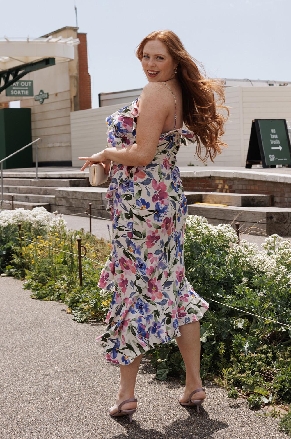 Person in a floral patterned sundress with ruffled hem walking outdoors while holding a cream clutch bag.