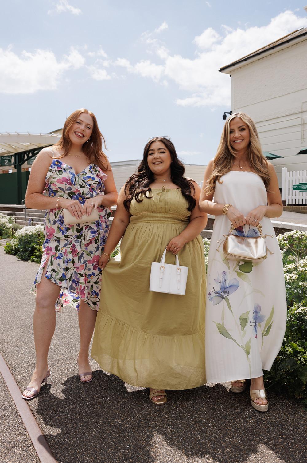 Three women wearing summer dresses posing outdoors. Left wears floral print, middle in gold maxi dress holds white handbag, right in white floral dress carries metallic bag.