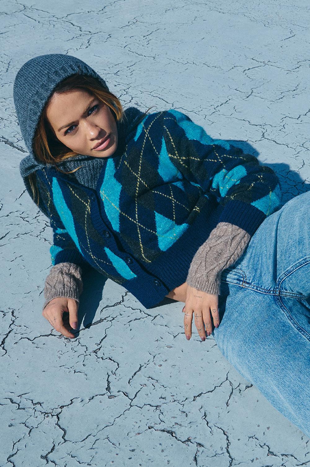 Ein Model in blauem Cardigan mit Argyle-Muster, grauer Beanie und heller Jeans sitzt auf einem ausgetrockneten Wüstenboden.