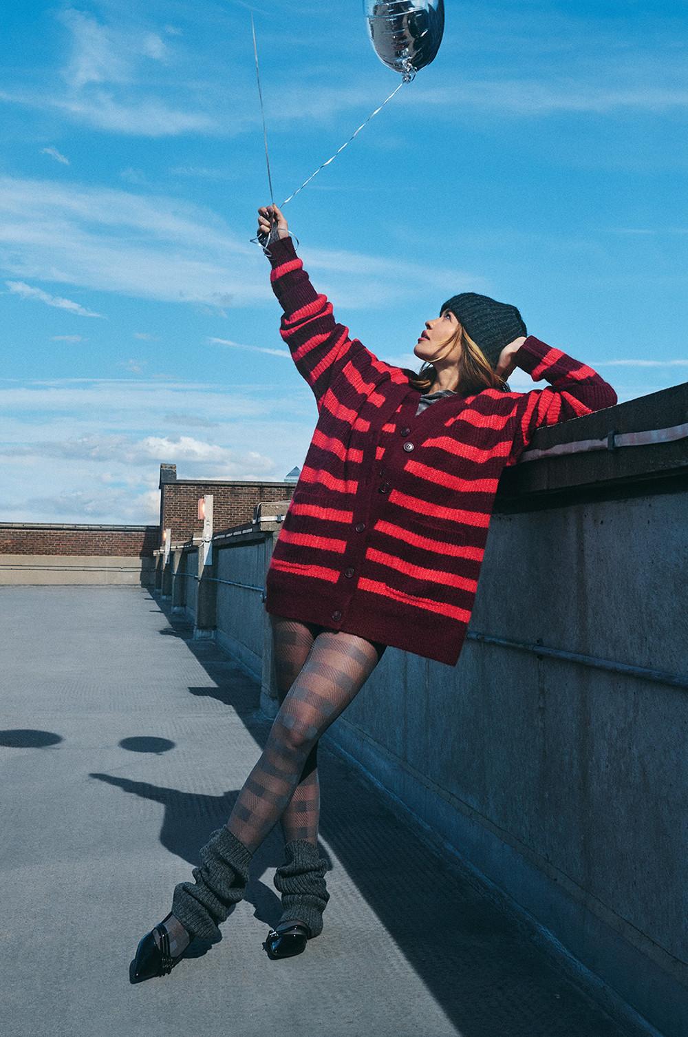 Model in einem rot gestreiften Cardigan mit schwarzer Beanie, das auf einem Dach einen silberfarbenen Ballon hält.