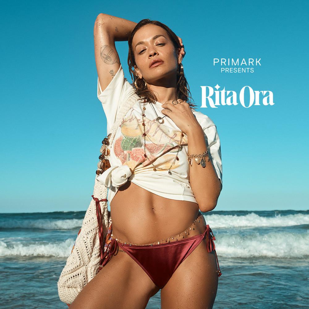 Rita Ora luce una camiseta blanca extragrande anudada, con estampado gráfico, bisutería y una braguita de bikini en tejido metalizado