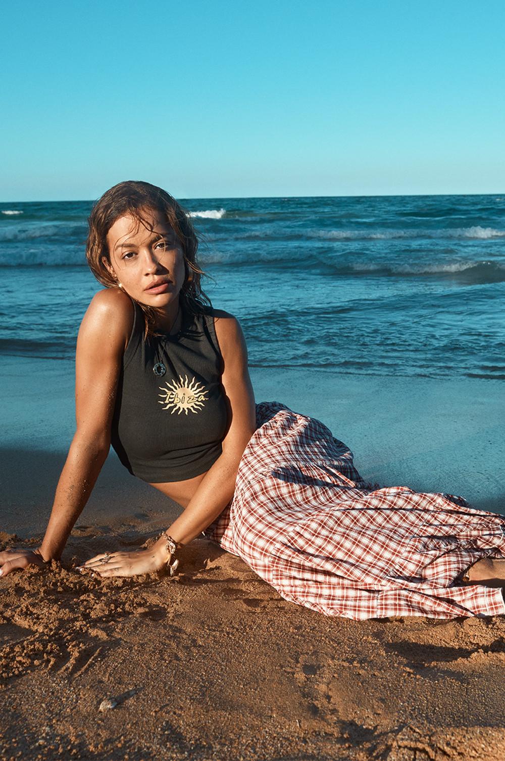 Rita Ora má na sebe červenú kockovanú vrstvenú maxi sukňu a čierne skrátené tielko s potlačou „Ibiza“ v strede
