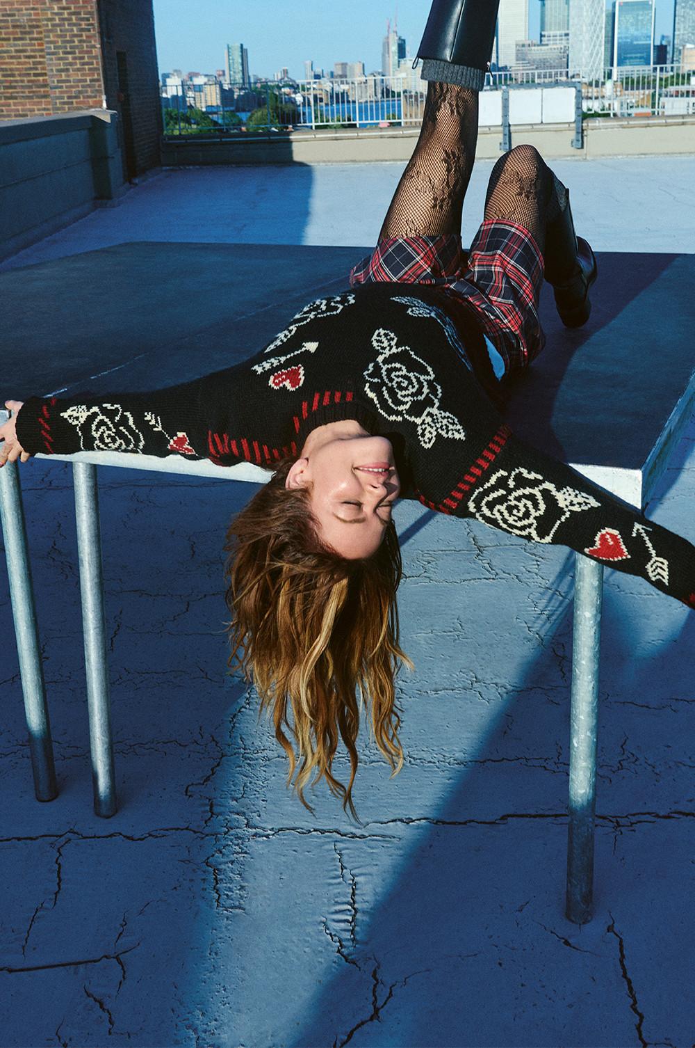 Personne portant un pull noir en maille à motif floral, une jupe à carreaux et des collants résille, sur un toit avec vue sur le paysage urbain.
