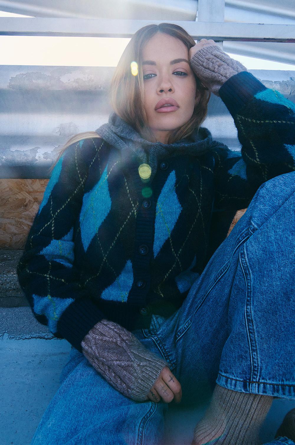 Personne portant un gilet à losanges bleu et noir, un jean en denim et des gants gris en tricot sans doigts.