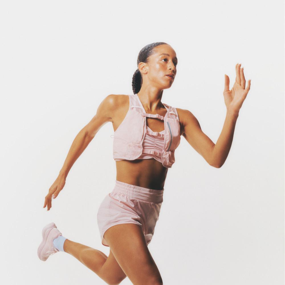 Persona su sfondo bianco che corre indossando un outfit sportivo rosa con top corto, pantaloncini e scarpe da ginnastica abbinate.