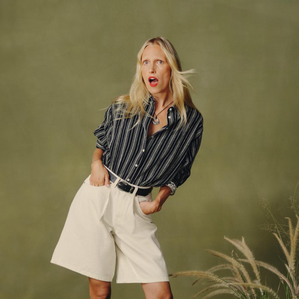 Modelo con camisa azul marino de raya diplomática, pantalones cortos blancos y cinturón negro, en una pose espontánea.