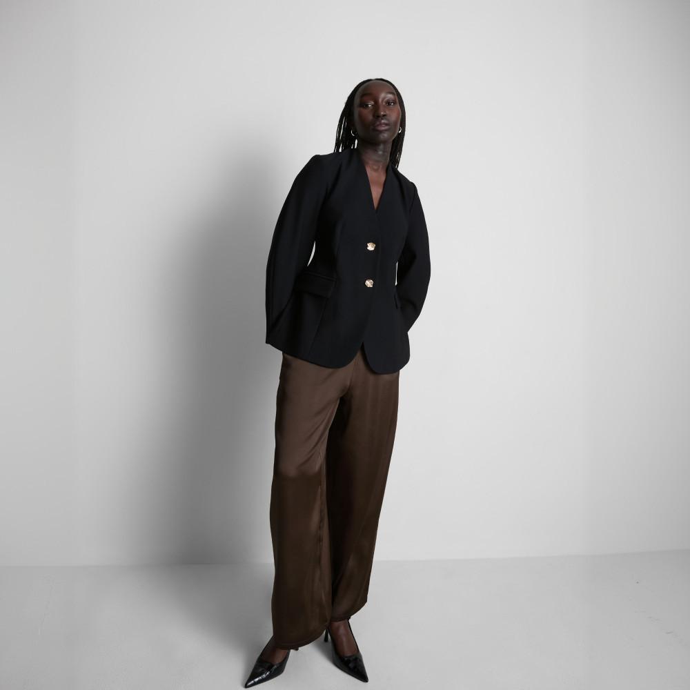 Modelo con blazer negra de vestir con botones plateados y pantalones marrones de pernera ancha.