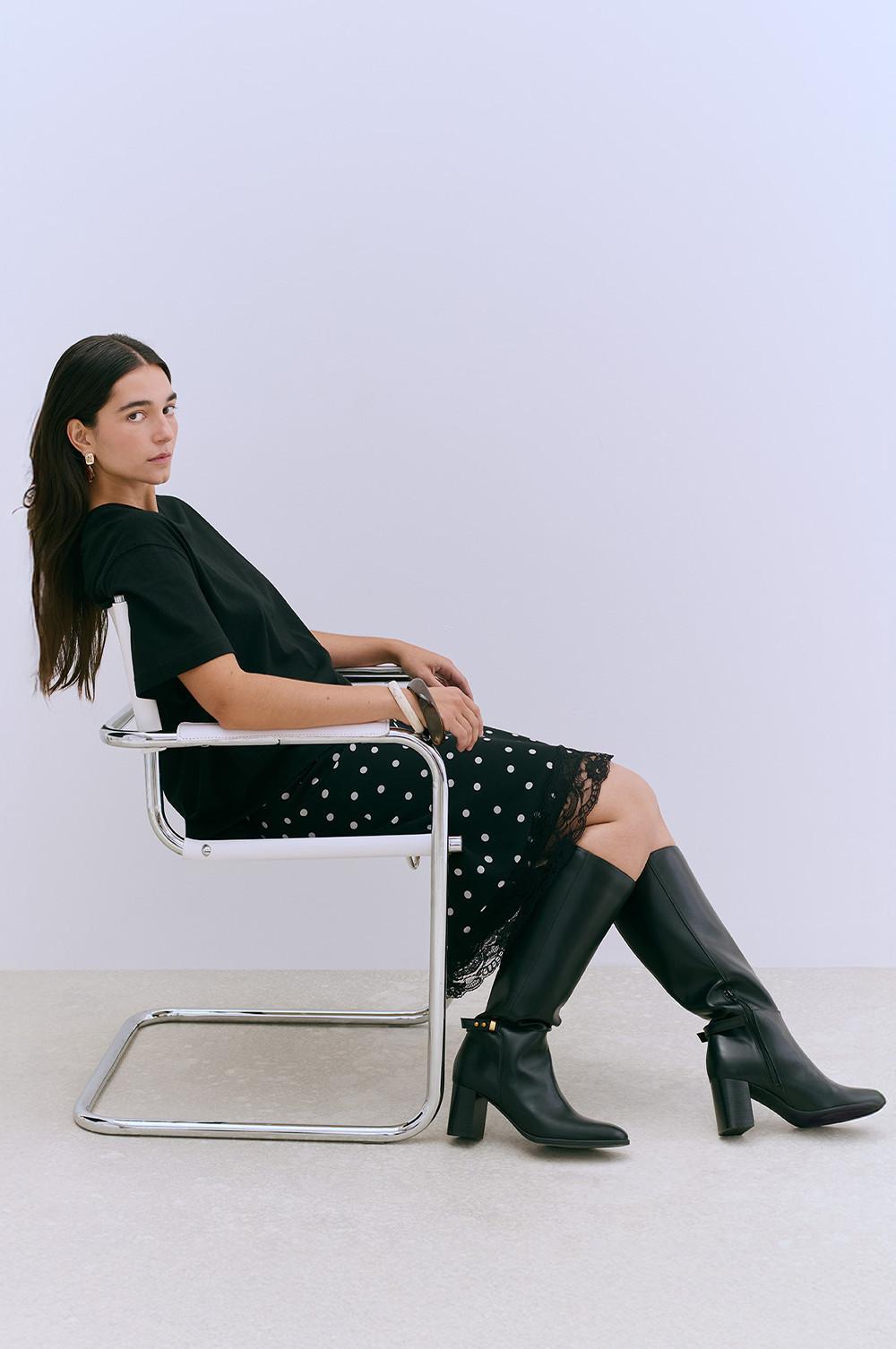 Person wearing black top, polka dot skirt with lace trim and knee-high leather boots, seated in a chrome chair.