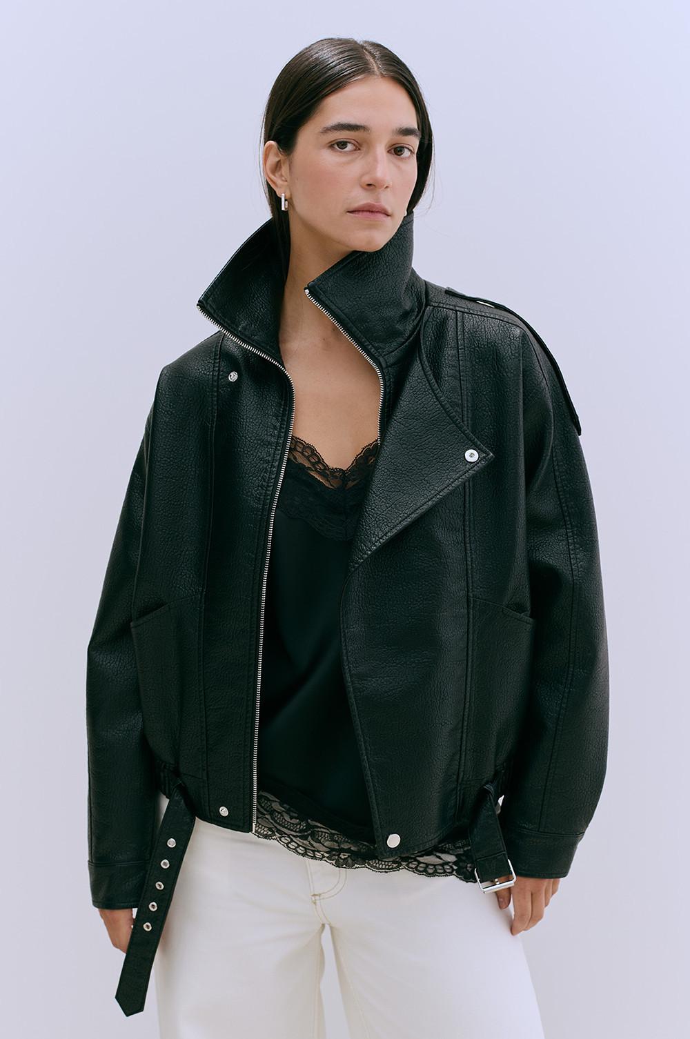 Oversized black leather jacket with high collar and belt detail, worn over lace-trimmed top with white trousers.