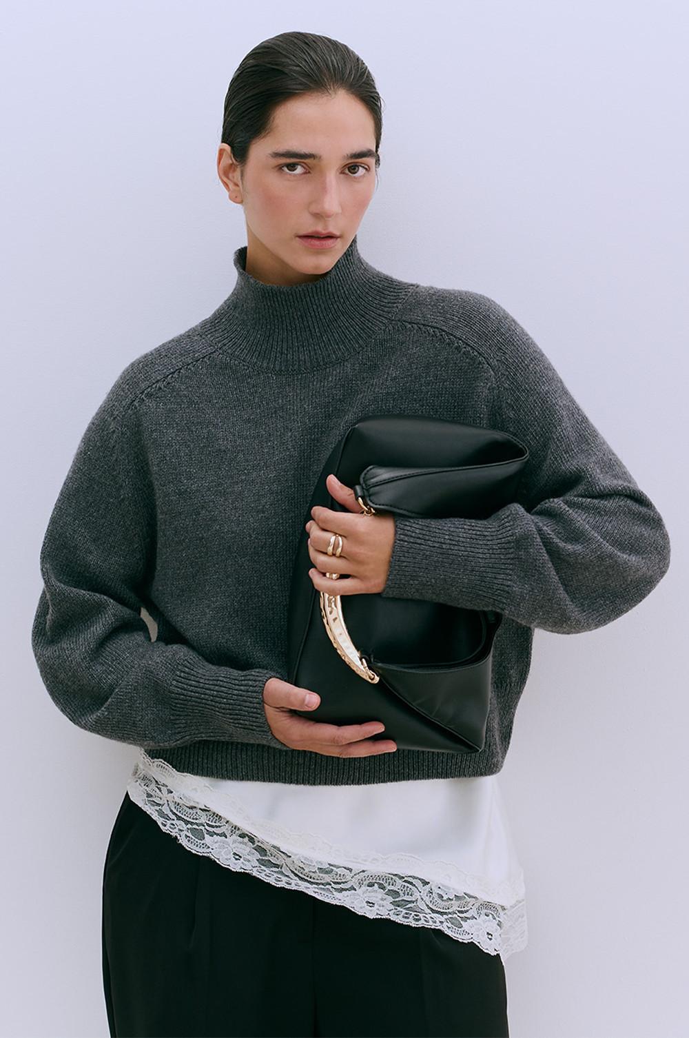 Person wearing grey turtleneck jumper with black clutch bag, white lace-trimmed top and dark trousers.