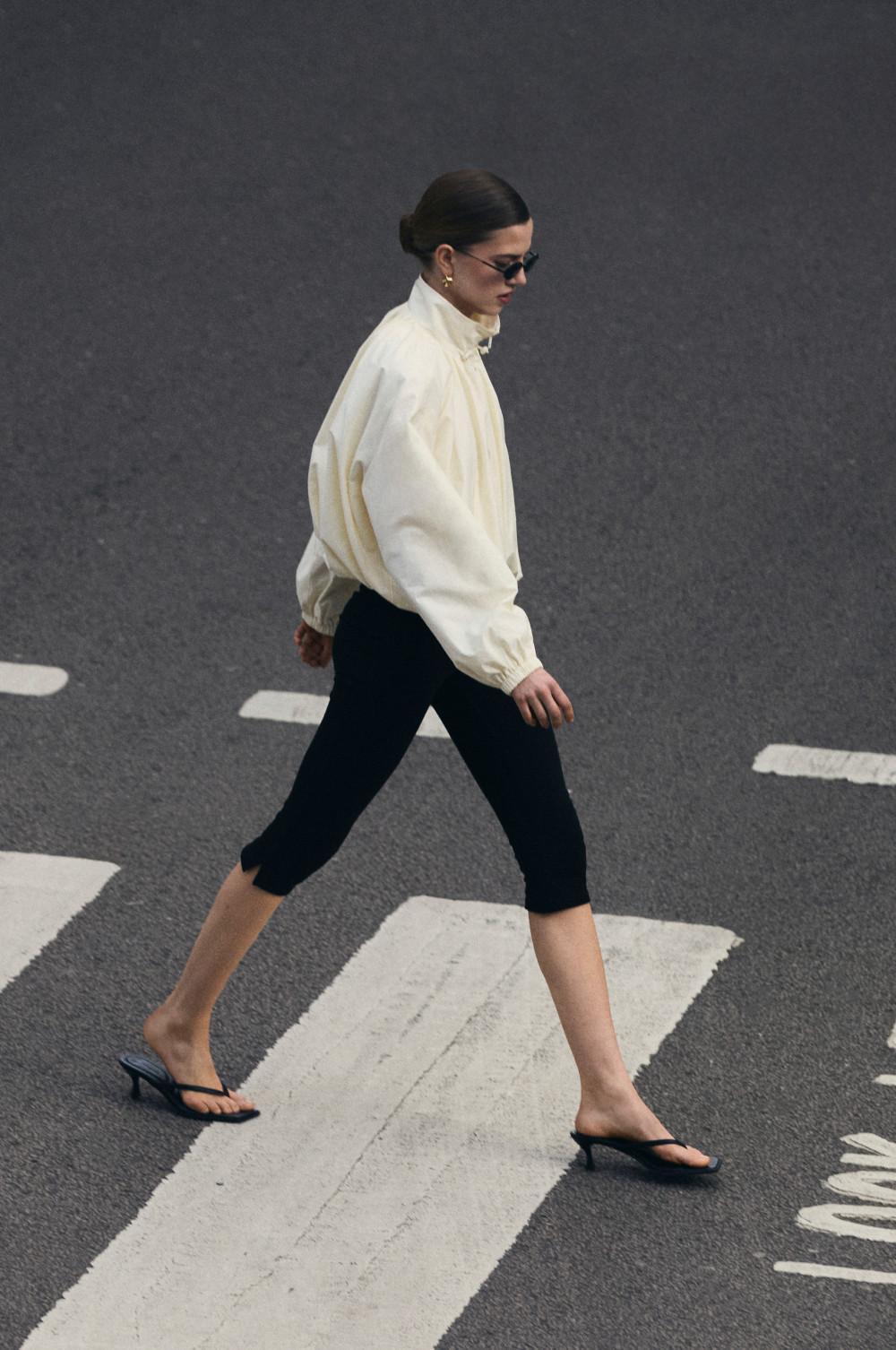 Woman walking across zebra crossing wearing cream oversized shirt, black cropped trousers and black flip-flops.