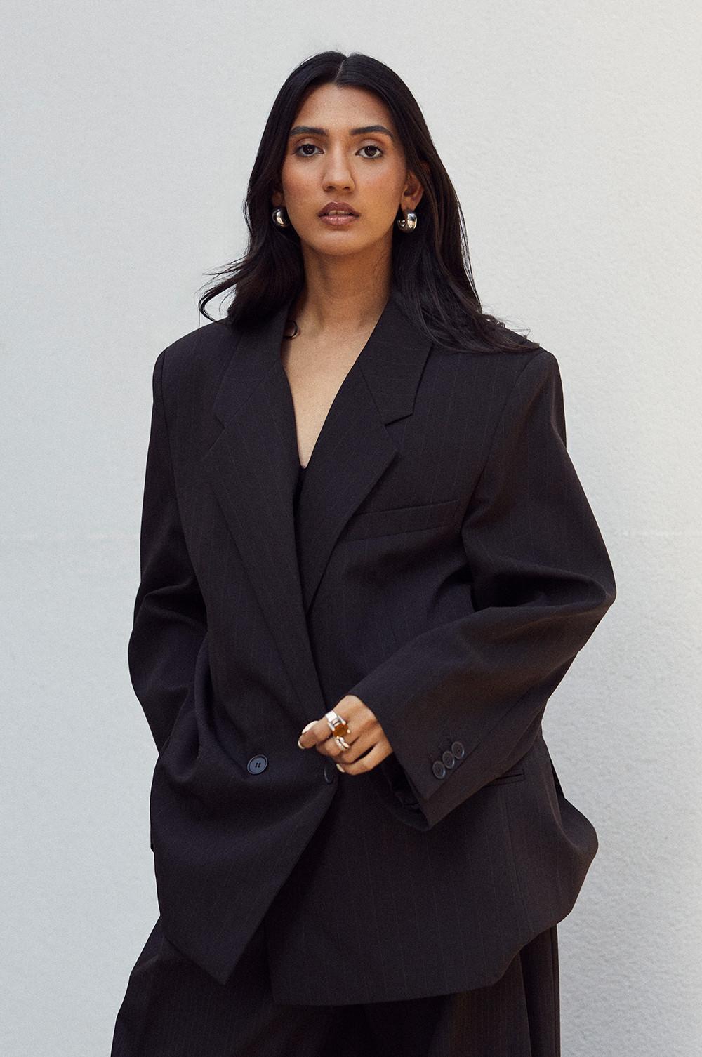 Sapna wearing an oversized black blazer with gold hoop earrings and statement rings against a white background.