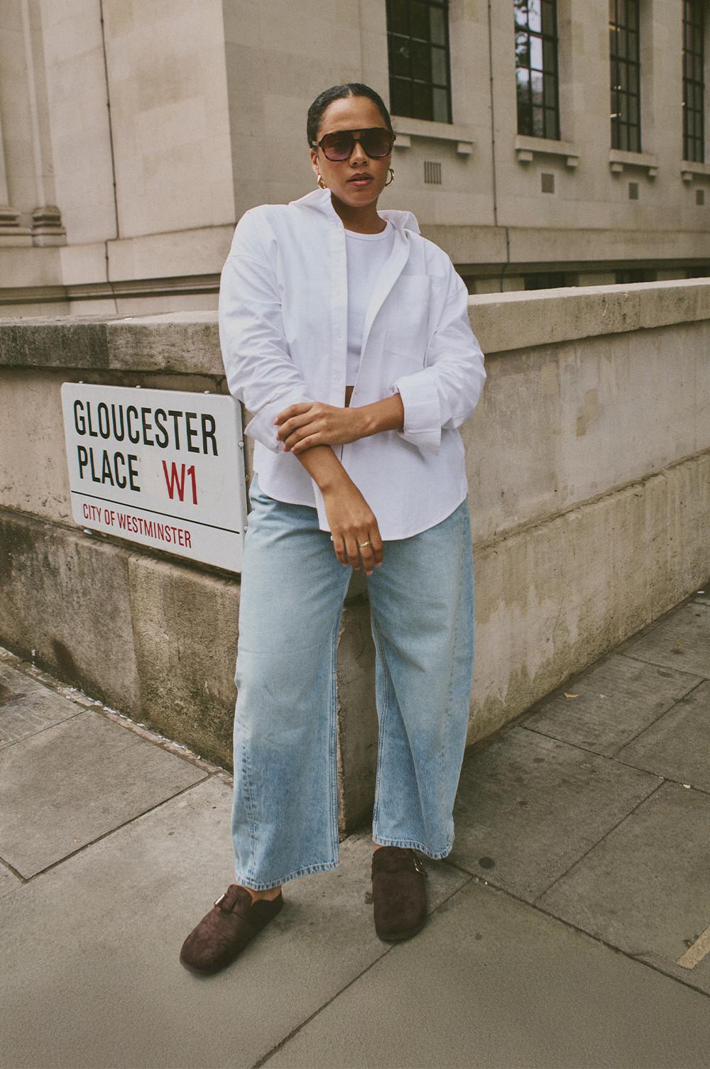 Person wearing white shirt, light blue wide-leg jeans and brown shoes standing near Gloucester Place sign in London.