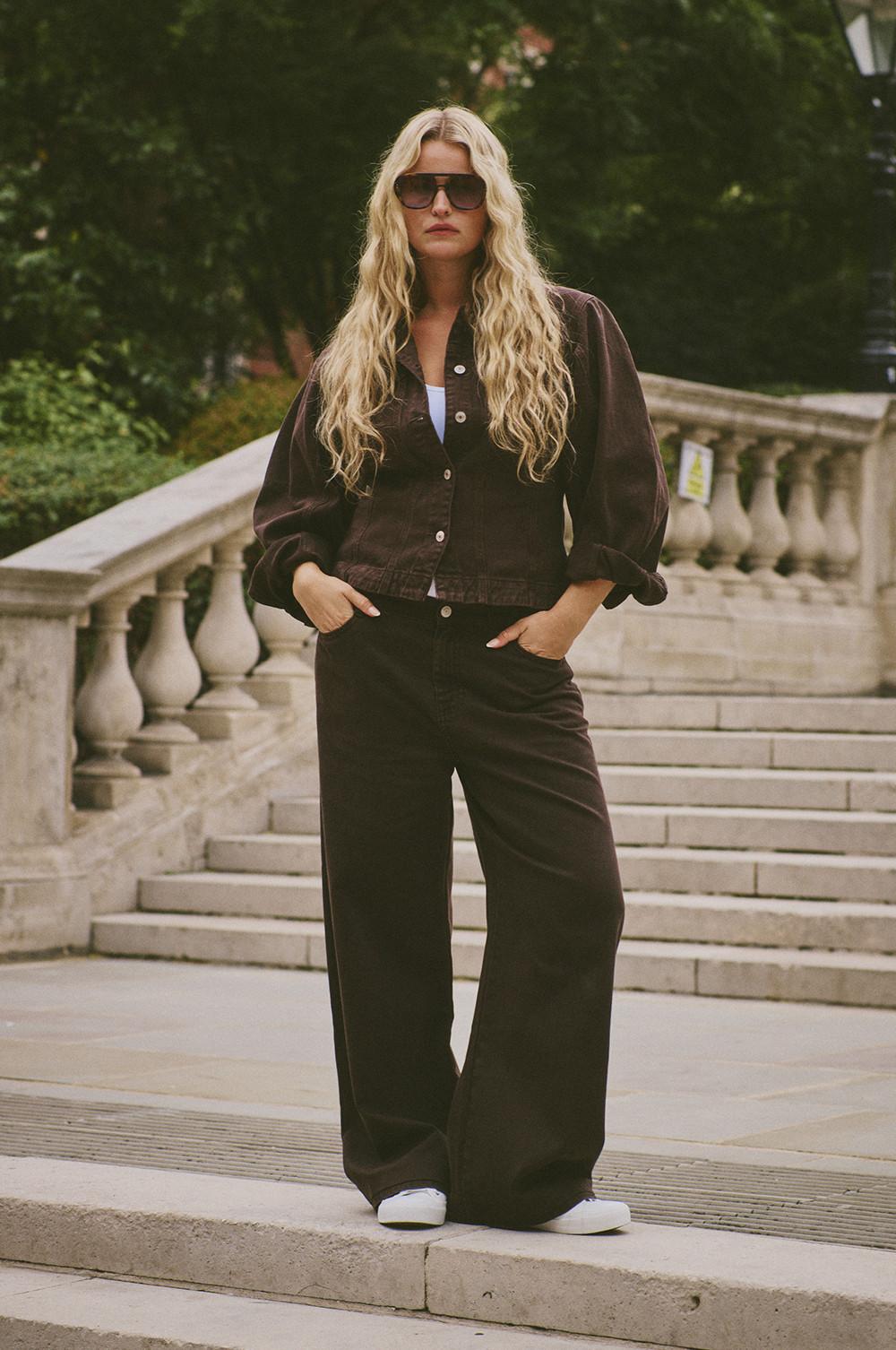 Person wearing brown denim jacket and wide-leg trousers with white trainers and sunglasses on stone steps.