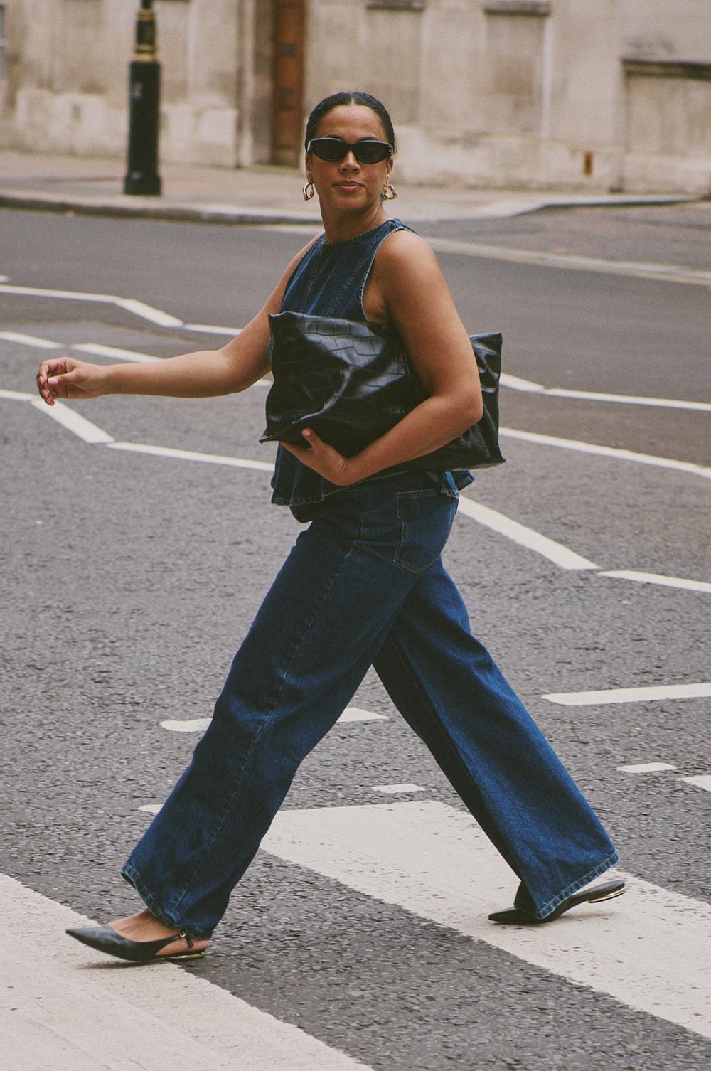 Person crossing street wearing denim outfit, sunglasses and carrying a black leather clutch bag.