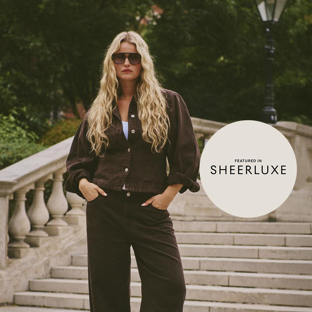 Person wearing brown jacket and trousers with sunglasses standing on stone steps, featured in SHEERLUXE.