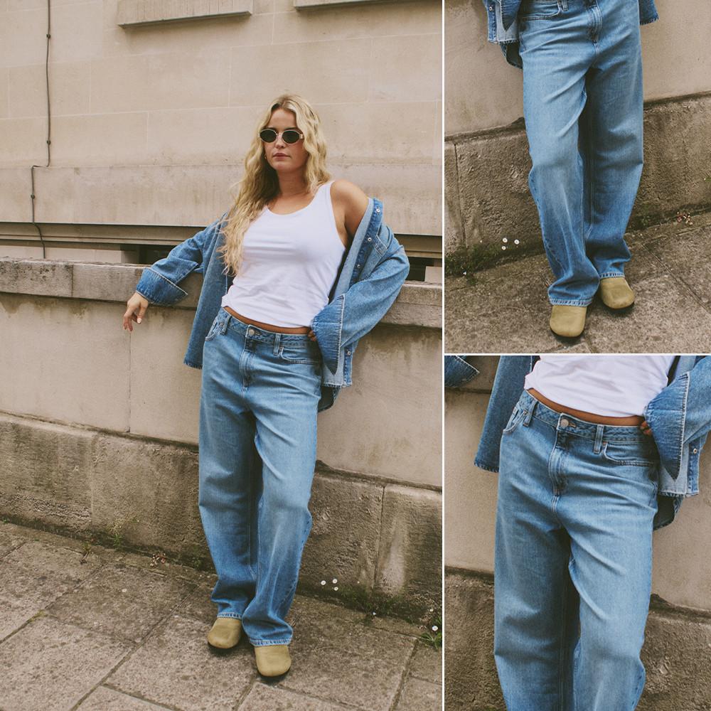 Persona che indossa una canotta bianca, una giacca di jeans e jeans dal taglio morbido con scarpe beige davanti a un muro di pietra.