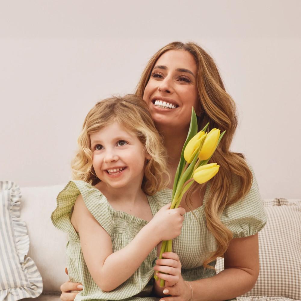 A smiling child holding yellow tulips while sitting with an adult in matching green gingham outfits on a sofa.