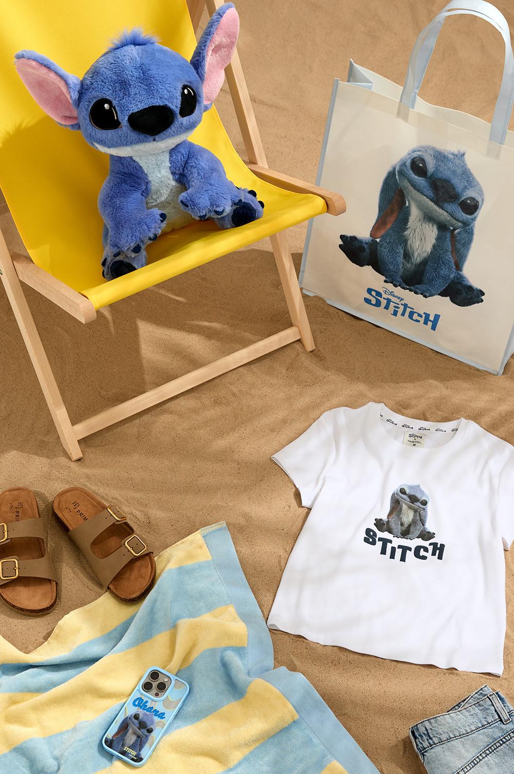 Stitch collection with a stitch plush toy, white baby graphic T-shirt, beach towel and shopping bag displayed in on a beach set.