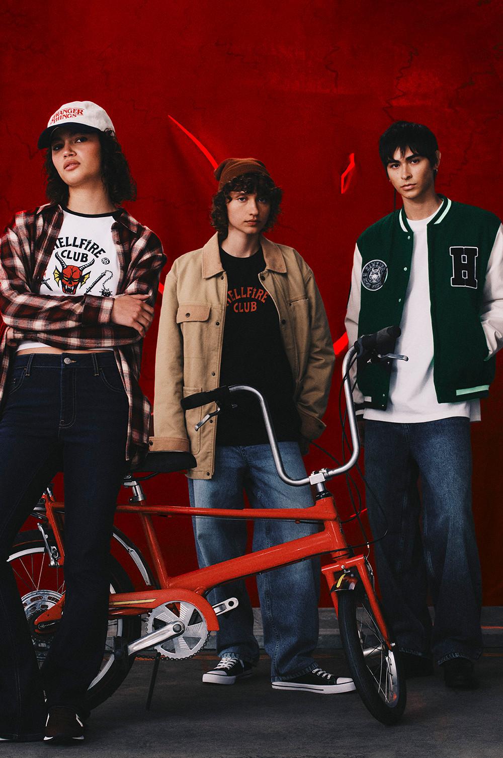 Three people wearing Hellfire Club merchandise posing with a red bicycle against a dark red background.