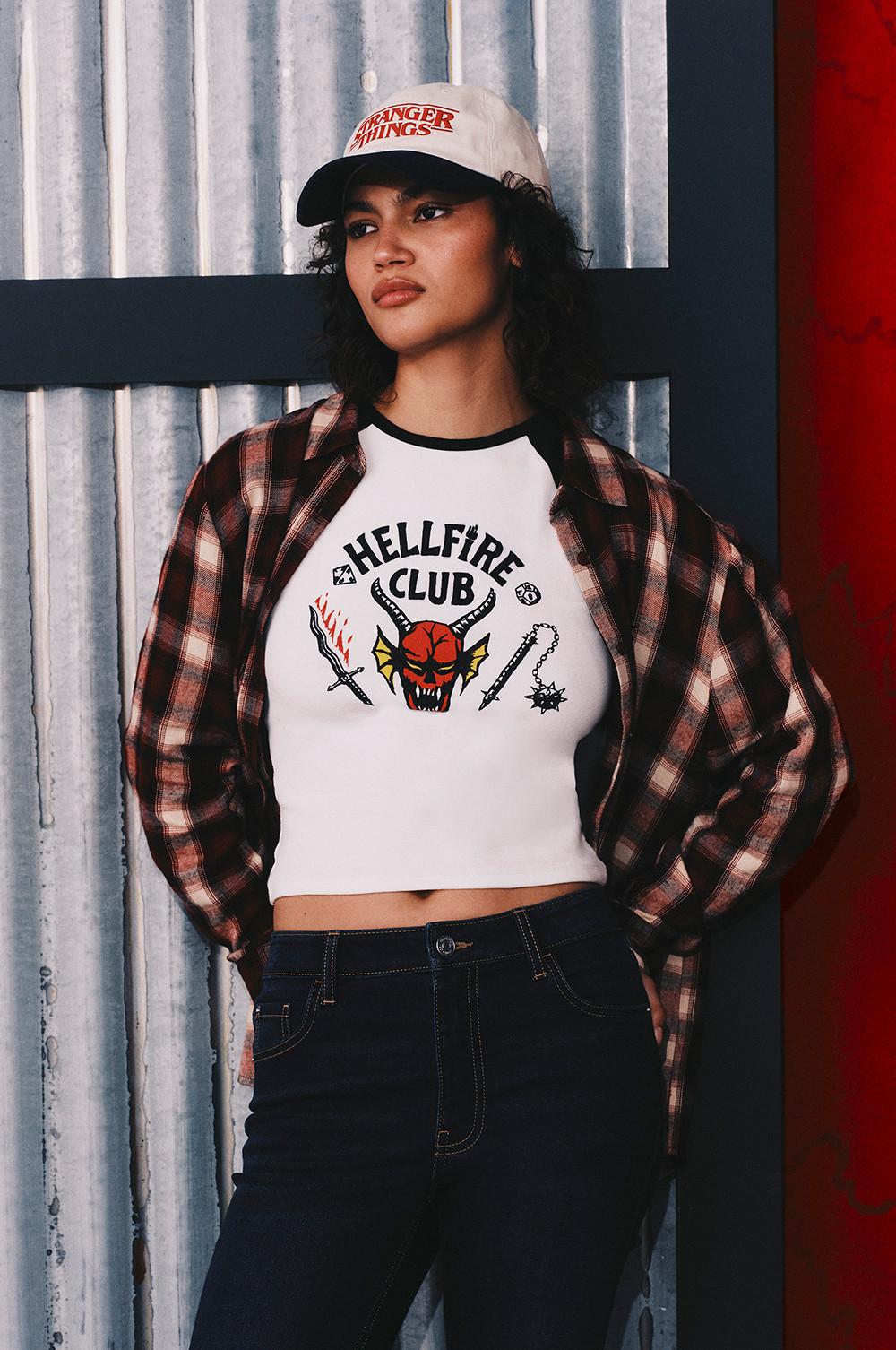 Person wearing Hellfire Club T-shirt with plaid shirt over top and Stranger Things cap against metal wall.