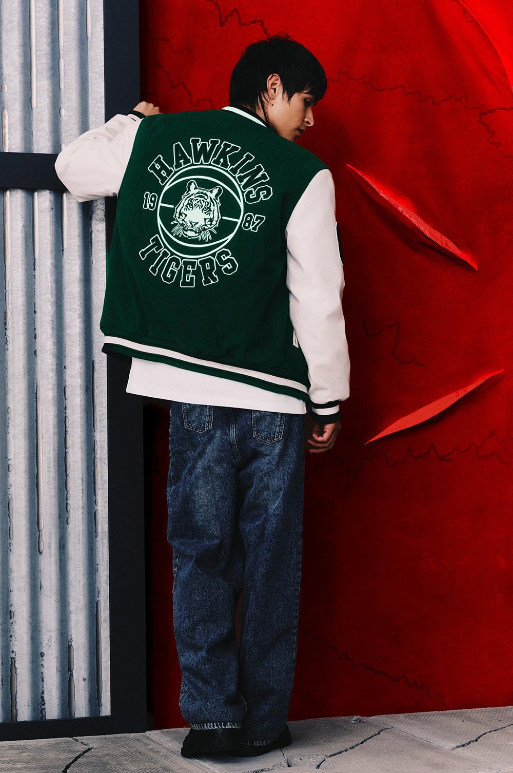 Person wearing green and white varsity jacket with Hawkins Tigers logo and 1987 emblem, paired with blue jeans.