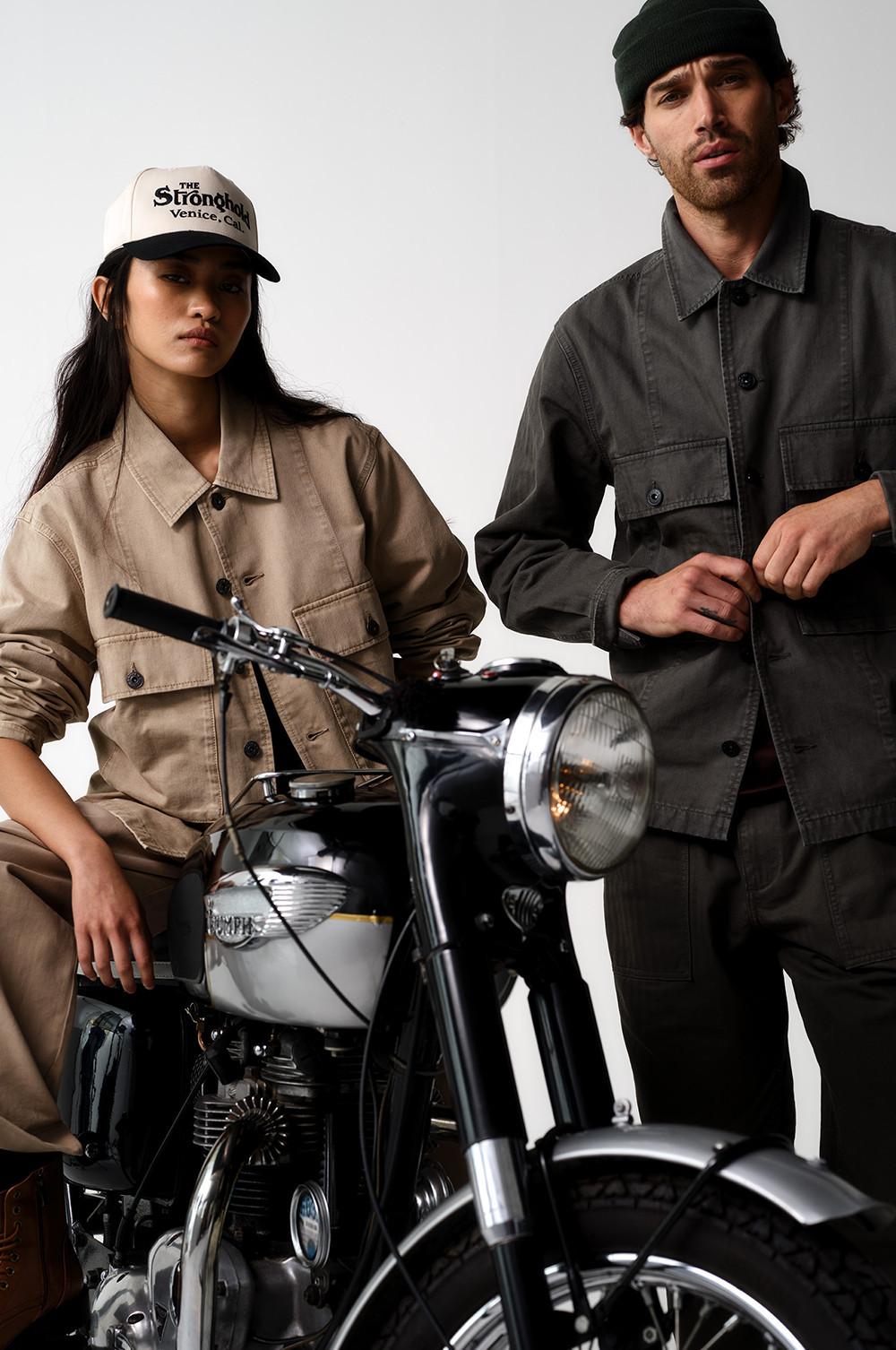 Due modelli con giacche utility in posa con una moto vintage; la modella indossa un cappellino "The Stronghold Venice".