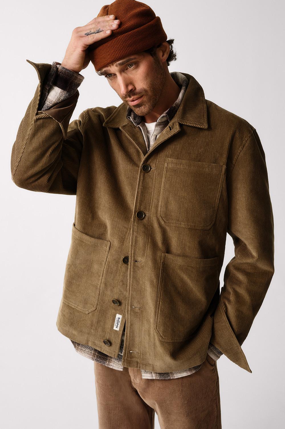 Person wearing brown corduroy jacket with multiple pockets, layered over a plaid shirt and topped with an orange beanie hat.