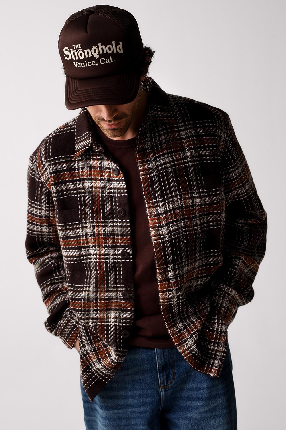 Person wearing brown Stronghold Venice trucker cap, plaid overshirt, brown T-shirt and denim jeans.