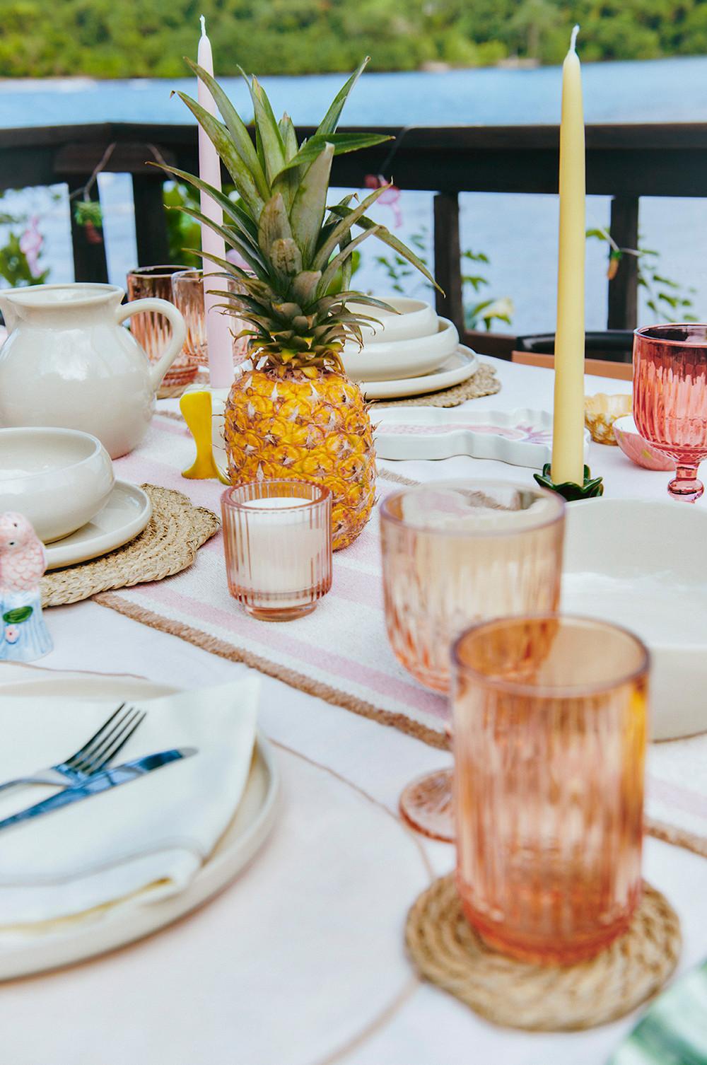 Table dressée avec de la vaisselle blanche et des sets de table tressés, un verre rose et un bol en forme d'ananas.