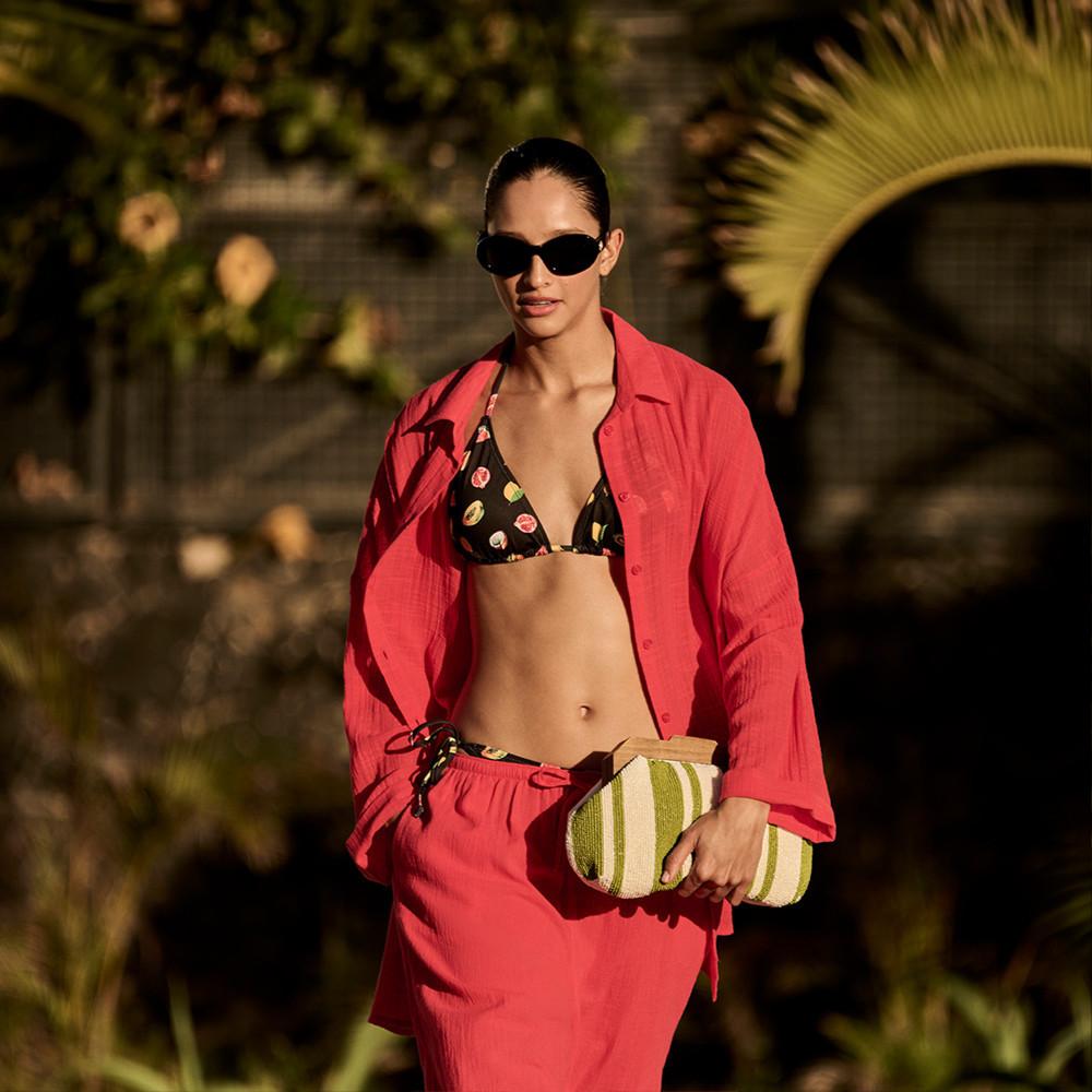 Modelo con traje de baño con cuello halter negro, gafas de sol y bisutería dorada, apoyada en una palmera.