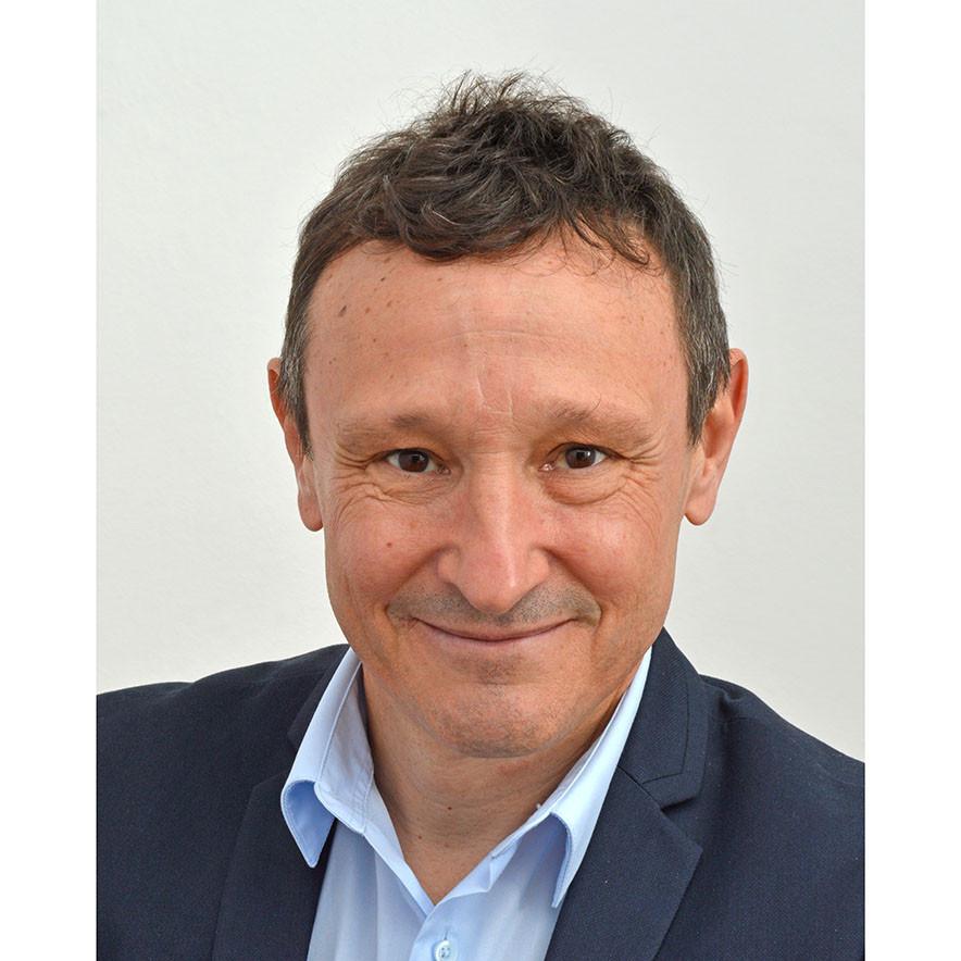 A smiling man in a dark blazer and light blue shirt against a plain background.