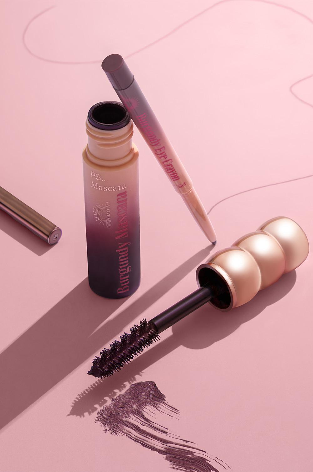 Mascara con esclusiva impugnatura, pastello per occhi bordeaux e campione di colore su una superficie rosa.