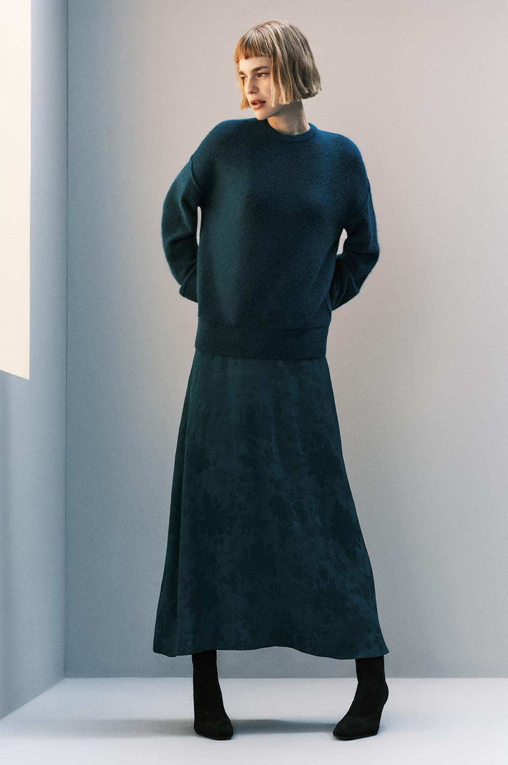 Person wearing teal jumper with matching long skirt and black ankle boots, standing against light wall.