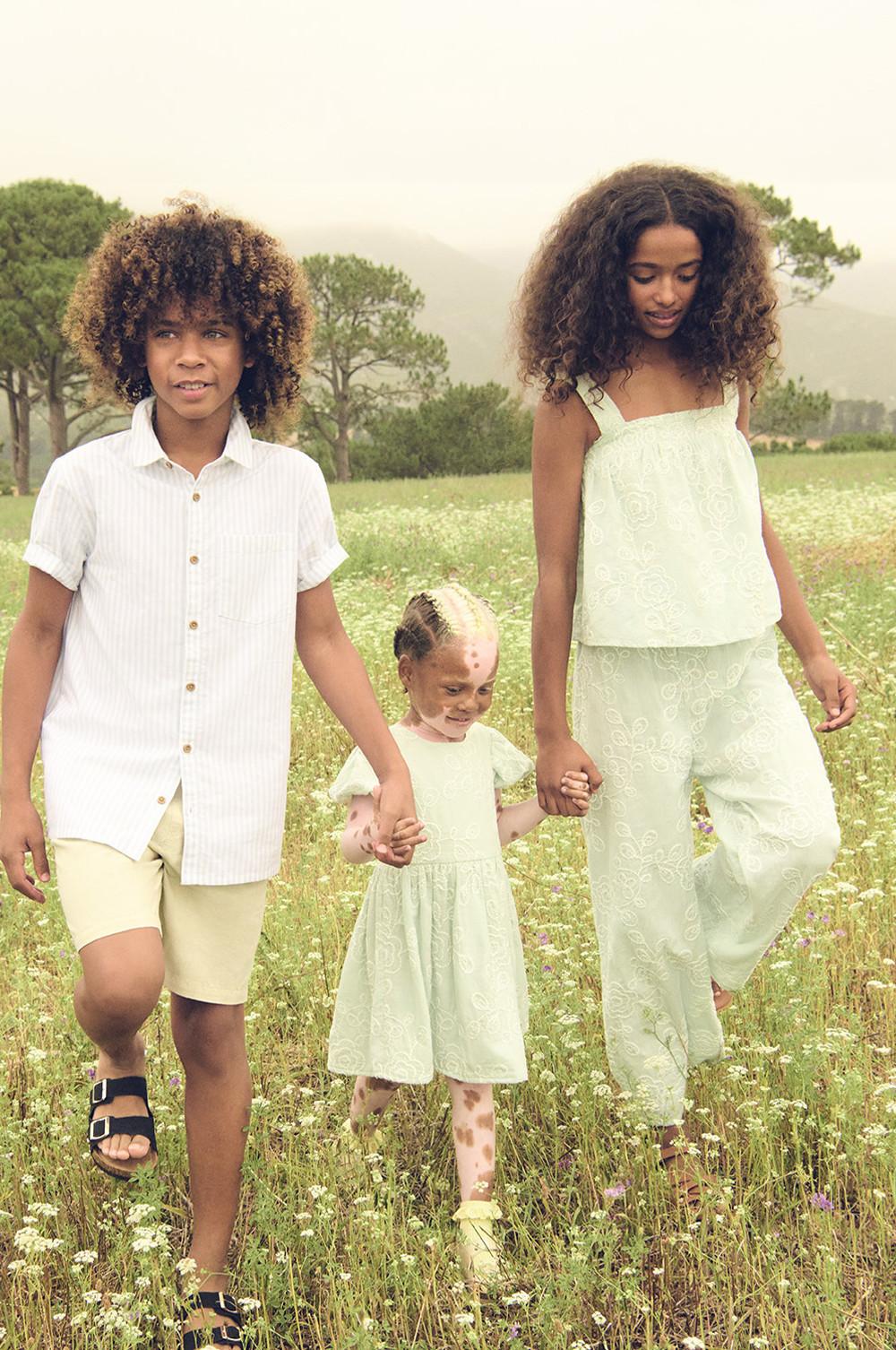 Family walking together holding hands through a summer meadow with tall grass and wildflowers wearing a white shirt, yellow shorts and black sandals, embroidered dress in pastel green and a co-ord in pastel green embroidery.
