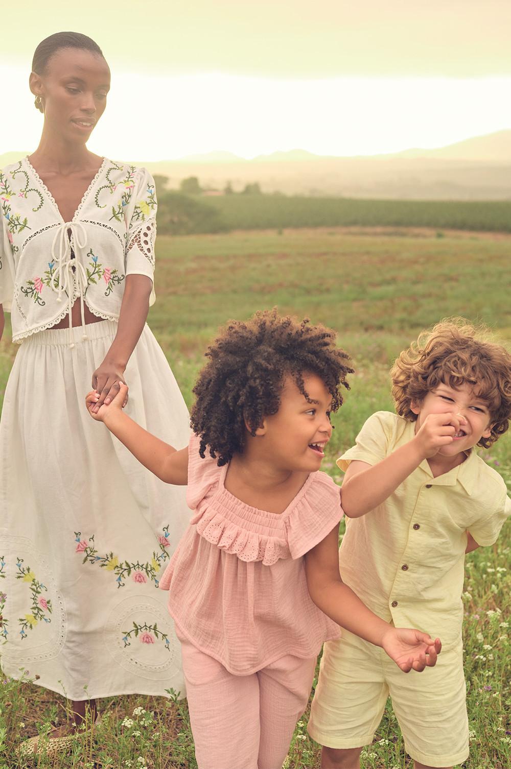 Three people in floral and ruffled dresses and shorts playing and running in a meadow at sunset.