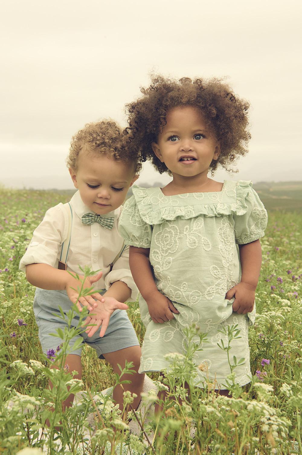 Two children playing together in a grassy field, both smiling and wearing light-colored dress, bow tie, shorts and shirt.