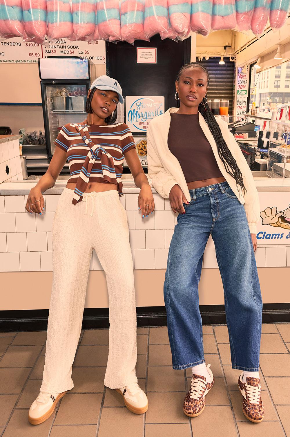 Two models posing in trendy outfits at a food counter with pink and blue cotton candy hanging above. Stylish casual wear with crop tops and wide leg trousers.