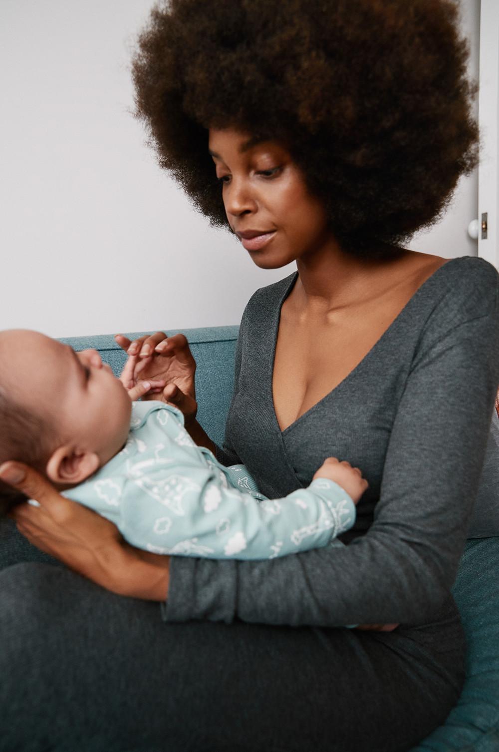 Model, das ihr Baby hält, sieht lächelnd nach unten und trägt ein graues Midikleid