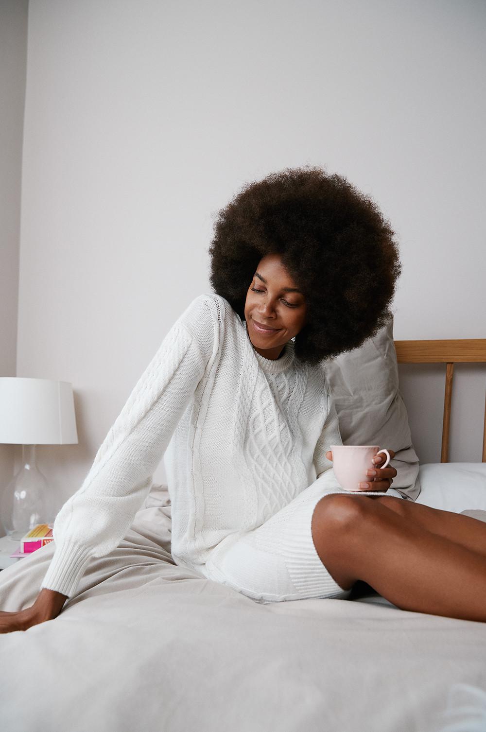 Modelo sentada en la cama con vestido de punto de ochos. Está sonriendo y sujeta una taza rosa.