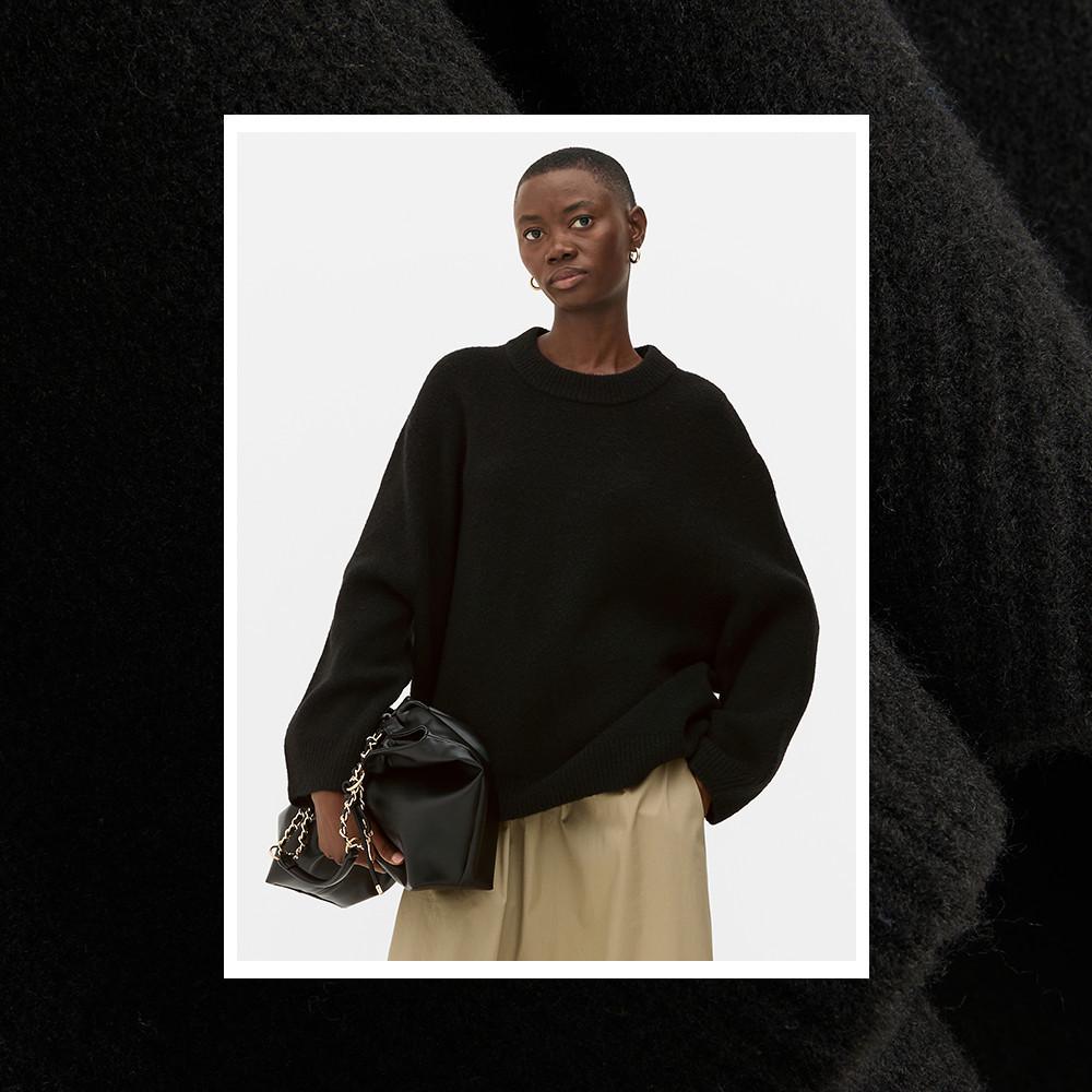 Person wearing a black oversized sweater with beige trousers, holding a black handbag with chain detail.