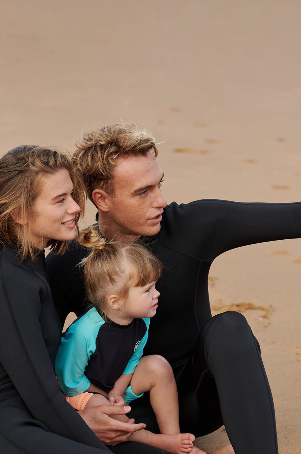 Family in wetsuits on beach; adults wear long black ones, child wears short