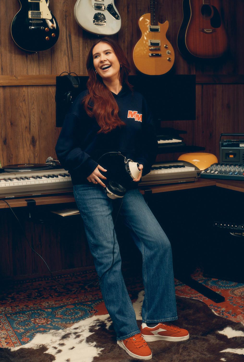 Person in music studio wearing navy hoodie and jeans, holding headphones with guitars displayed on wall.