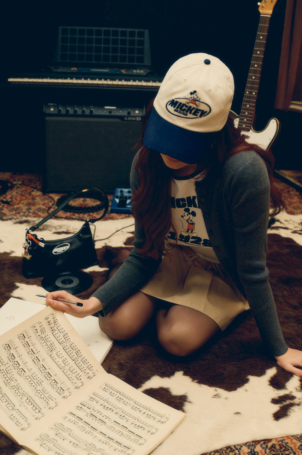 Person wearing Mickey Mouse cap and t-shirt writing music notes in a home studio with keyboard and guitar.