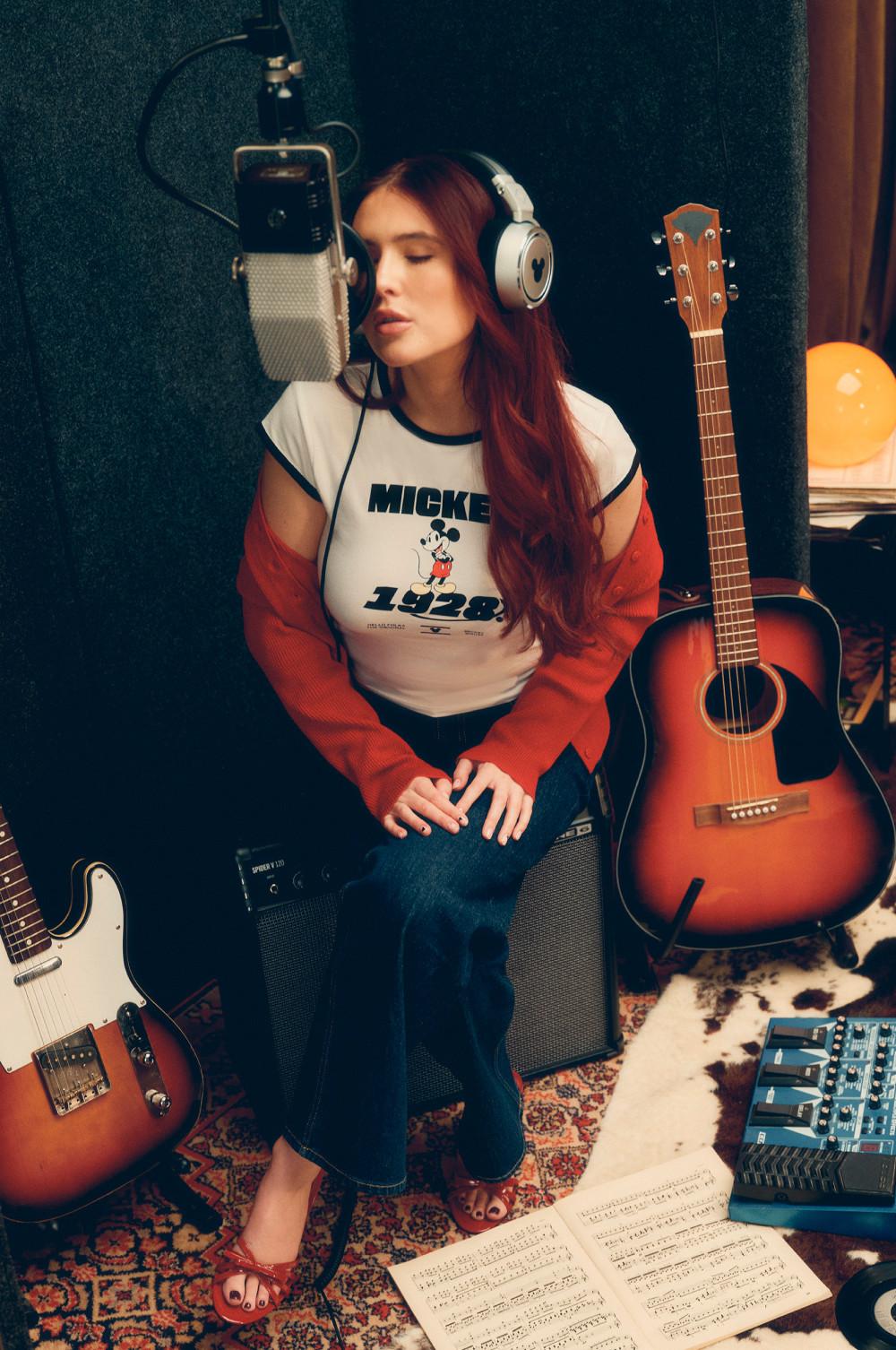Person recording music in studio wearing Mickey Mouse t-shirt, surrounded by acoustic guitar and equipment.