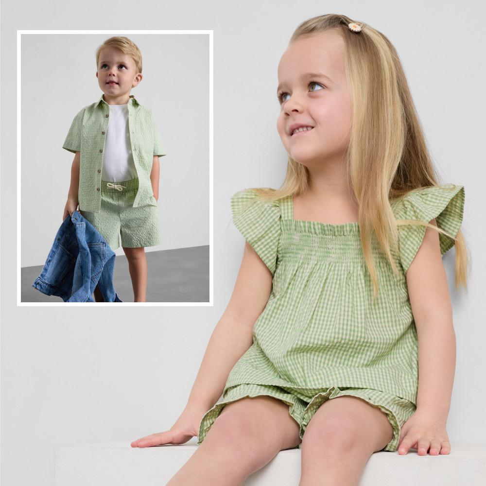 Sitzendes und lächelndes Mädchen in grünem Kleid mit Vichykaros und Rüschenärmeln mit abgestimmten Jungen-Outfit.