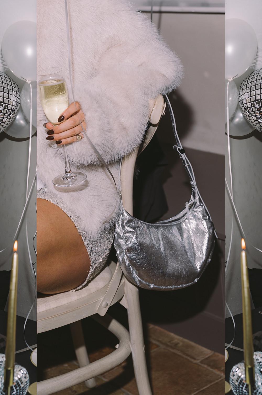 Modella che indossa un cappotto in pelliccia bianca e tiene in mano un bicchiere di champagne. Una borsetta argentata è appesa a una sedia.