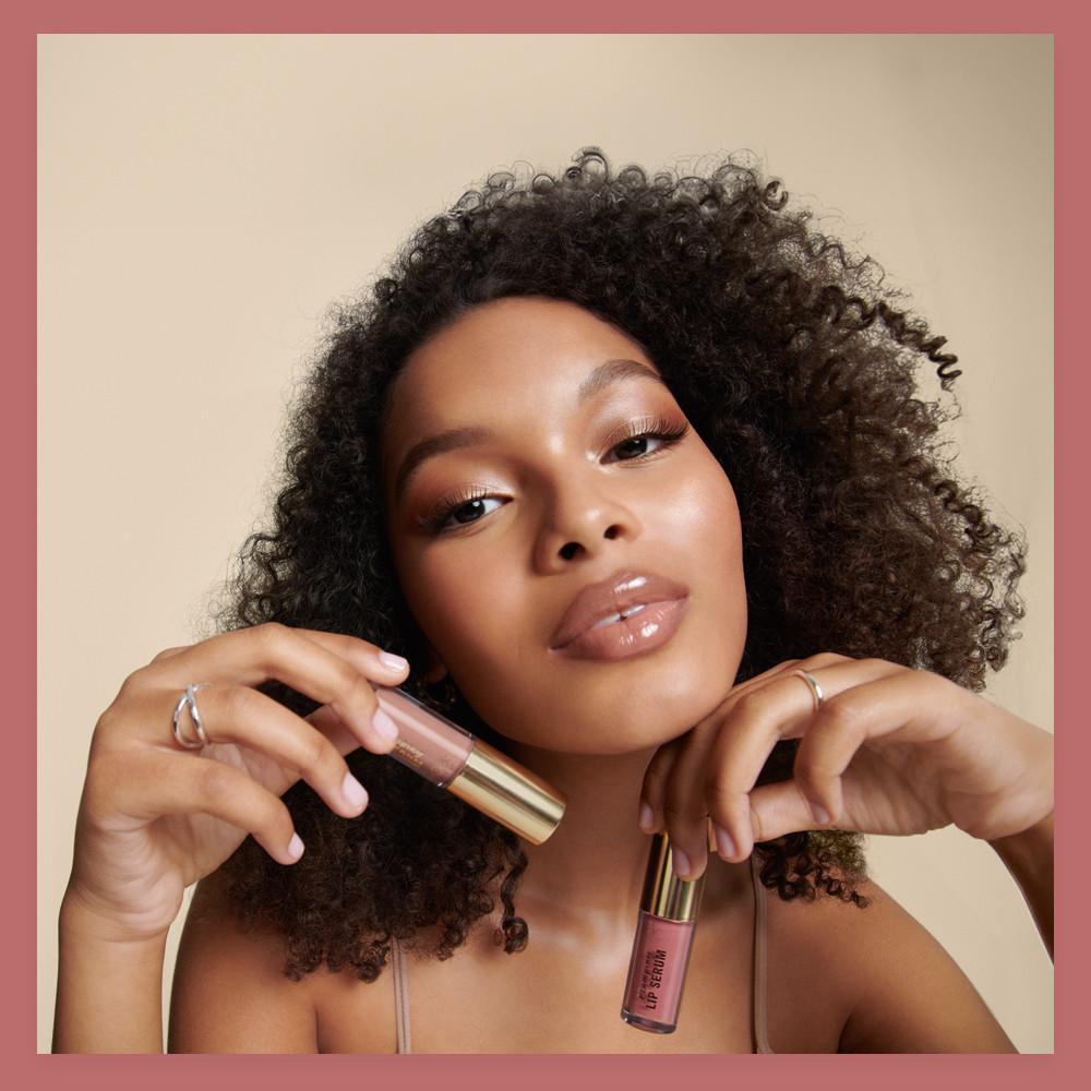 Beauty model holding two nude-toned lip products with gold-toned caps against beige backdrop, wearing glossy makeup look.